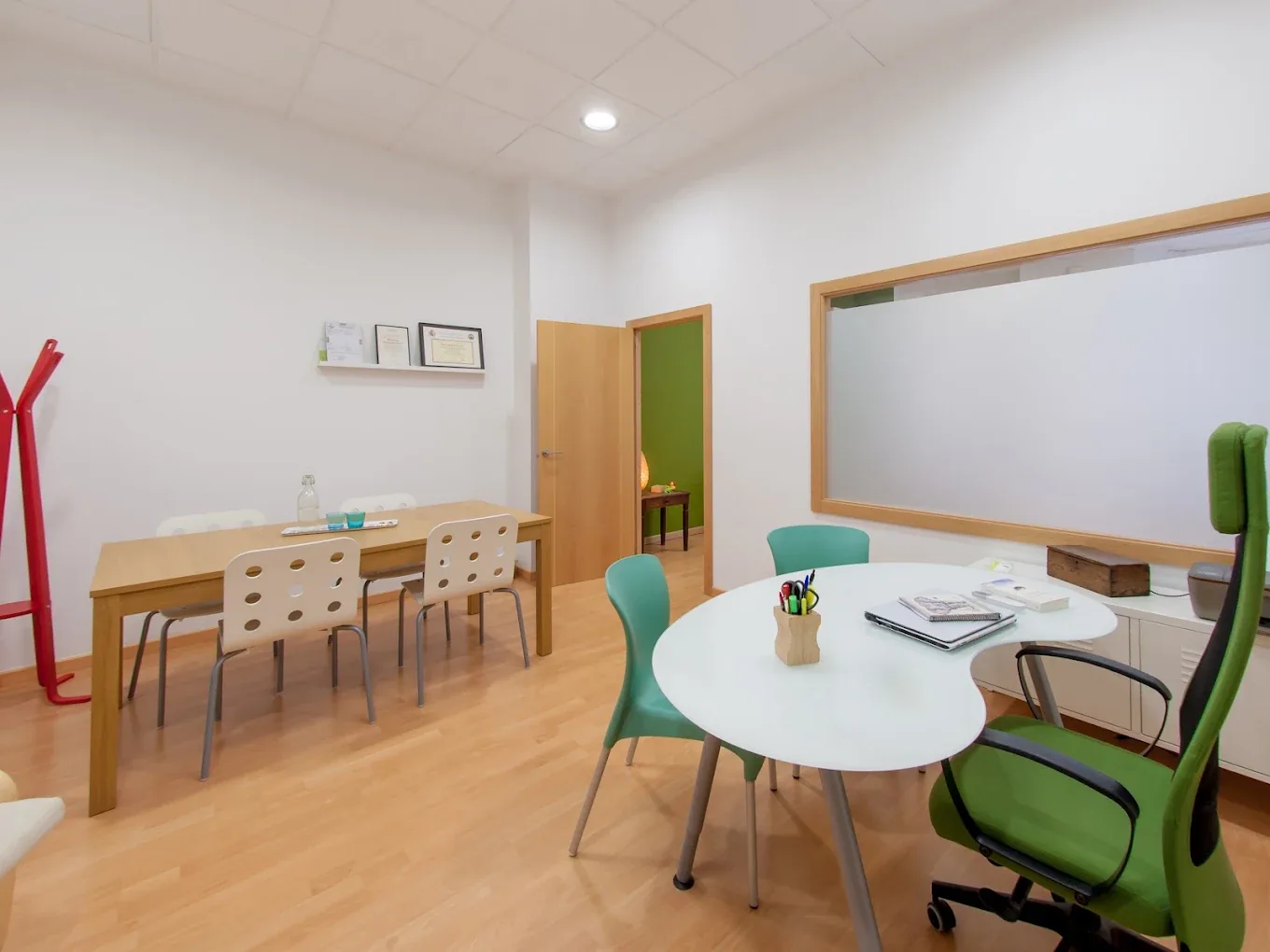 Despacho de la consulta con mesa redonda, sillas verdes y una mesa de trabajo junto a la ventana