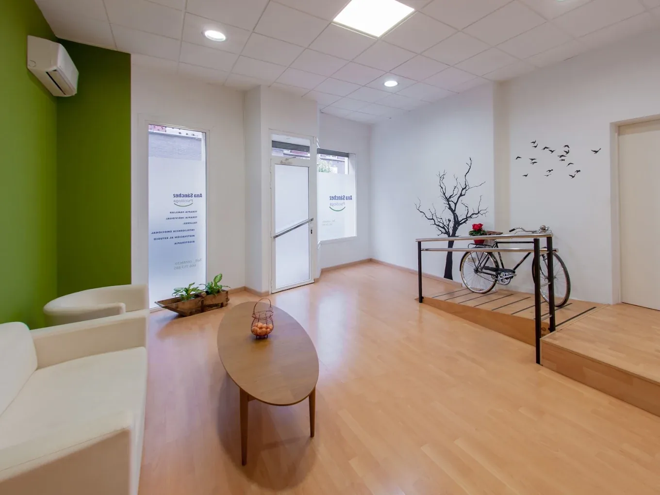 Sala de espera de la consulta: sofá blanco, pared verde, suelo de madera y un vinilo de un árbol en la pared